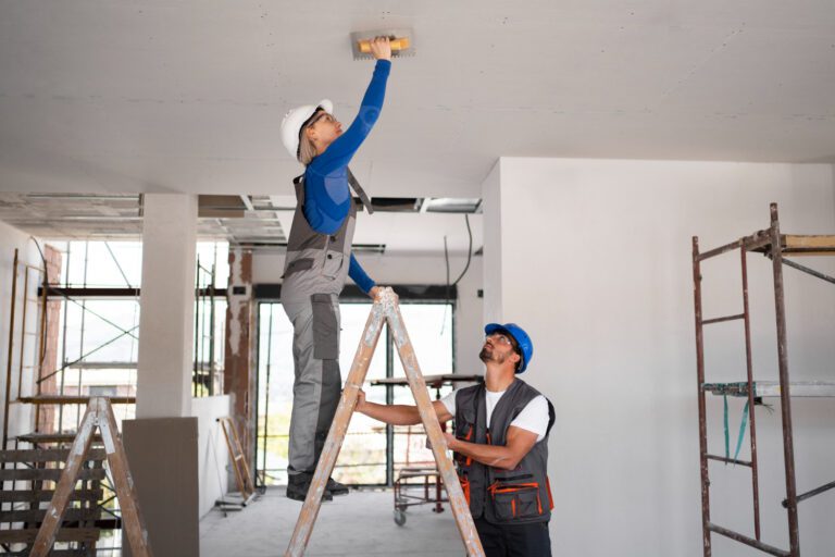 Construction workers wearing safety helmets and vests, one standing on a ladder sanding a ceiling while the other stabilizes the ladder inside a building under renovation.