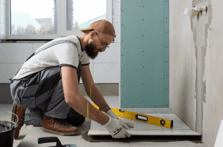 Man installing floor tiles in a bathroom remodel, using a level tool to ensure precision. He's wearing work gloves, overalls, and a backwards cap.