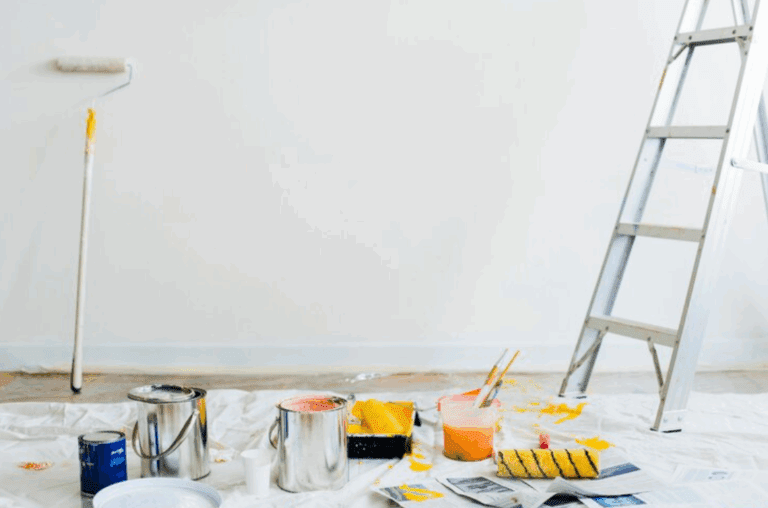 Room prepared for painting with drop cloth on the floor, open paint cans in yellow and orange, paint rollers, brushes, and a metal ladder against a white wall.