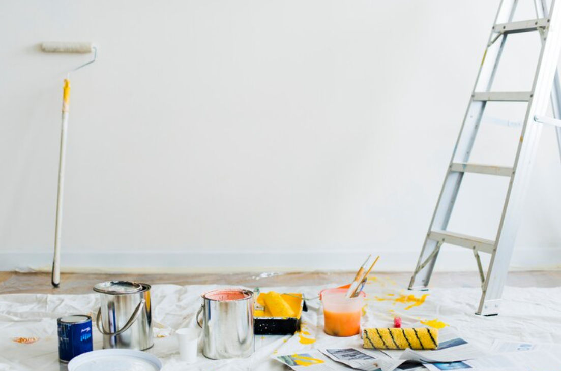 Room prepared for painting with drop cloth on the floor, open paint cans in yellow and orange, paint rollers, brushes, and a metal ladder against a white wall.