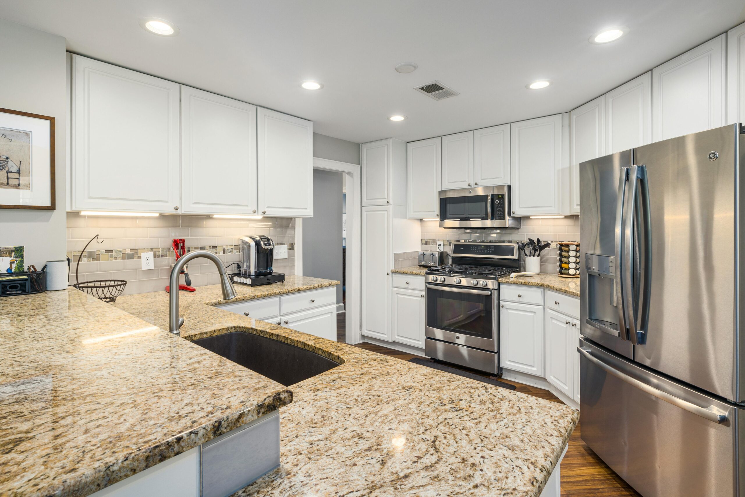 Modern U-shaped kitchen with white shaker cabinets, stainless steel appliances, and granite countertops. The space features a built-in microwave, double-door refrigerator, and under-cabinet lighting, with a sleek gooseneck faucet over a large undermount sink and warm wooden flooring adding contrast.