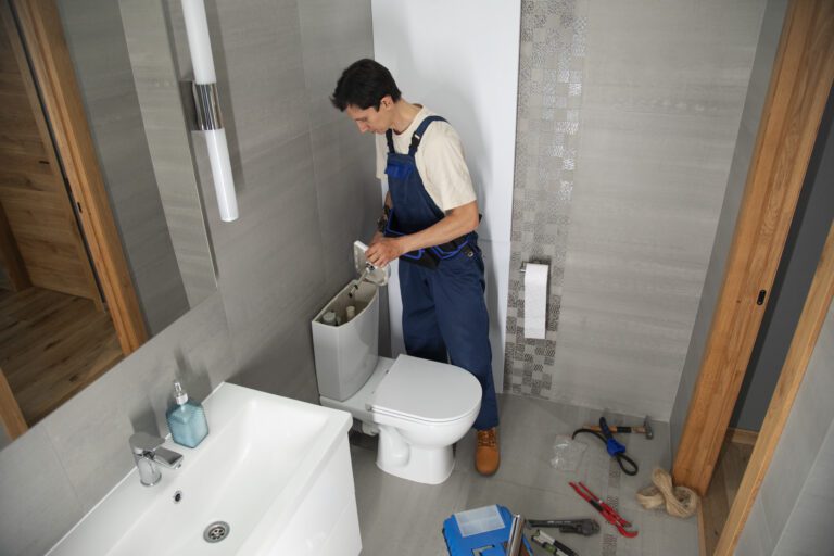 Professional plumber repairing a toilet tank in a modern bathroom with plumbing tools on the floor.