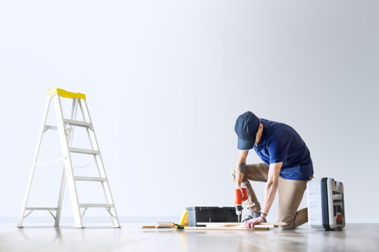 Handyman using a power drill on wooden boards in a bright room, with ladder and toolbox nearby during home renovation.