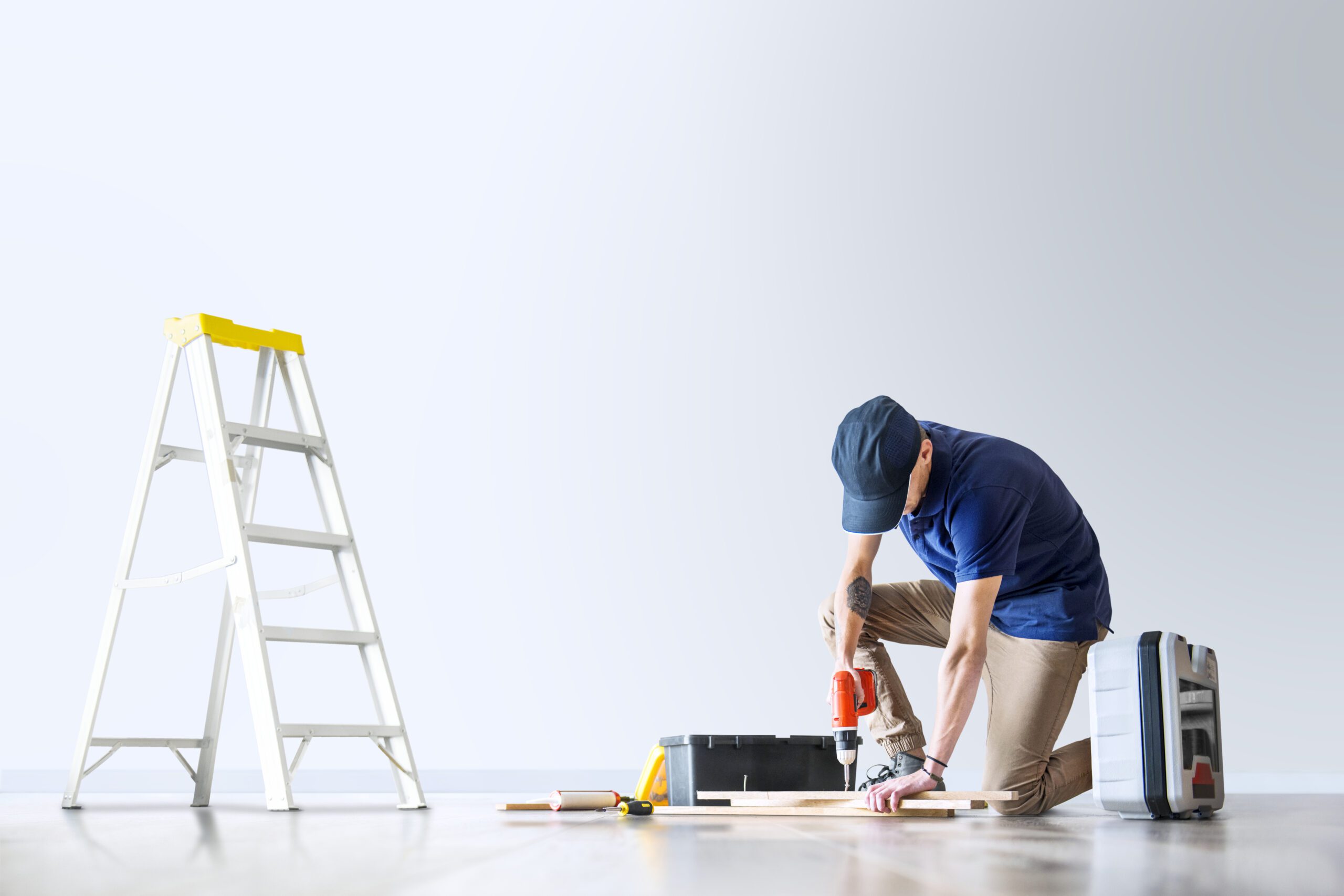 Handyman using a power drill on wooden boards in a bright room, with ladder and toolbox nearby during home renovation.
